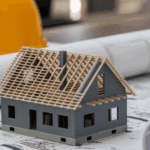 Image of a model house under construction, laying on a blueprint with a hardhat visible.